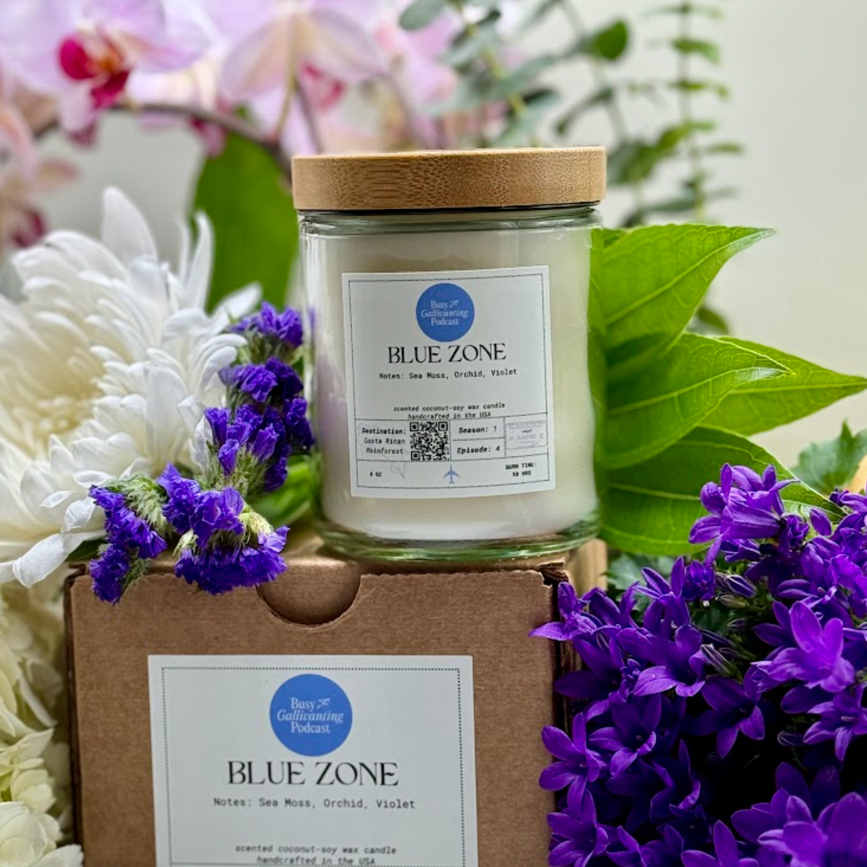 Blue Zone candle on top of candle box with label, surrounded by white, pink and floral flowers— hydrangeas, plumeria and orchids