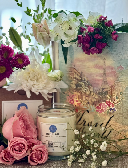 Busy Gal candle surrounded by pink roses, white lillies, baby's breath, and "travel the world" map book